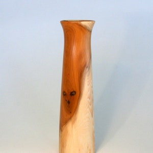May include: A tall, slender wooden vase with a natural wood finish. The vase is light brown in colour with darker brown streaks throughout. The vase is made from a single piece of wood and has a smooth, polished finish.