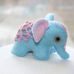 May include: A small blue elephant plush toy with pink crocheted details and a pink fabric ear. The elephant has a black button eye and is sitting on a white surface.