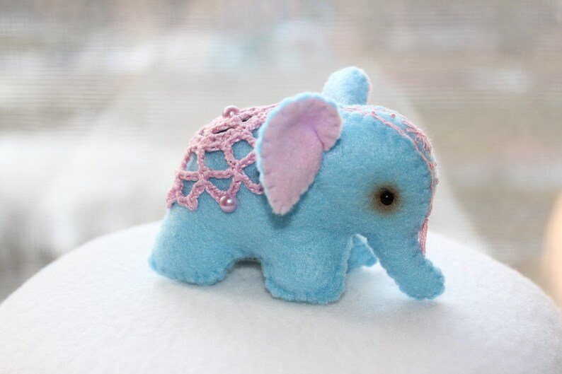 May include: A small blue elephant plush toy with pink crocheted details and a pink fabric ear. The elephant has a black button eye and is sitting on a white surface.