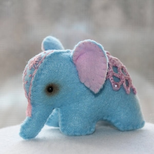 May include: A small blue felt elephant toy with pink felt ears and a pink crocheted trim. The elephant has a stitched-on eye.