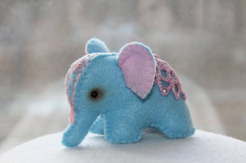 May include: A small blue felt elephant toy with pink felt ears and a pink crocheted trim. The elephant has a stitched-on eye.