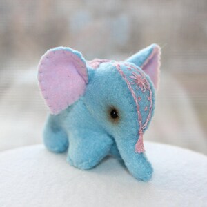 May include: A small blue plush elephant toy with pink ears and a pink embroidered design on its head. The elephant has a small pink tassel hanging from its head.