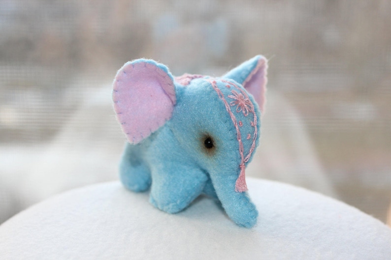 May include: A small blue plush elephant toy with pink ears and a pink embroidered design on its head. The elephant has a small pink tassel hanging from its head.
