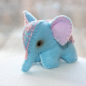May include: A light blue felt elephant toy with pink ears and embroidered details. The elephant has a single black eye and a pink and white tassel hanging from its trunk.