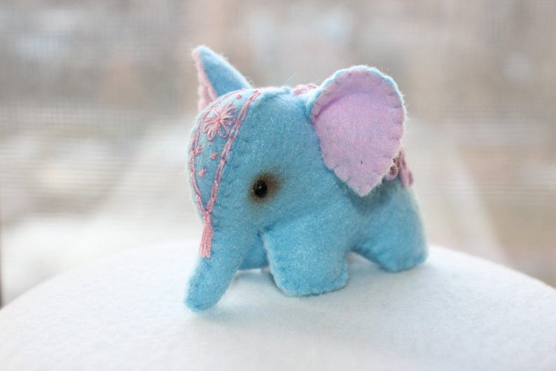May include: A light blue felt elephant toy with pink ears and embroidered details. The elephant has a single black eye and a pink and white tassel hanging from its trunk.