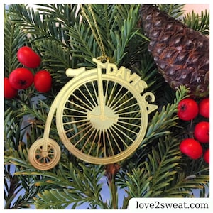 May include: Gold-colored metal ornament shaped like a bicycle with the name "Davis" written on the wheel. The ornament is hanging on a green pine branch.
