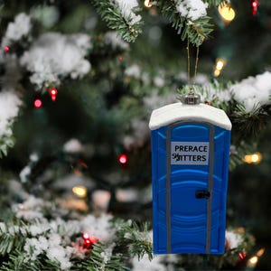 May include: A blue and white novelty Christmas ornament shaped like a portable toilet. The ornament has the words "PRERACE SHITTER" printed on the front. It hangs from a gold string against a snowy Christmas tree background.