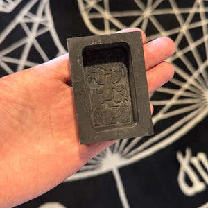May include: A dark grey rectangular mould featuring an embossed dragon design and the text "COBBLE 999 FINE". The mould is held in a hand, with a black and white patterned background.