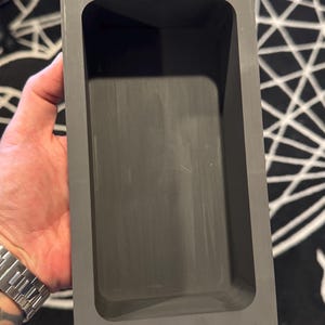 Puede incluir: Un recipiente rectangular gris oscuro con la parte superior abierta y esquinas redondeadas. El recipiente se sostiene en la mano de una persona, mostrando su forma y profundidad. La superficie tiene un acabado mate.