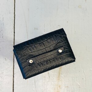 K&ouml;nnte beinhalten: Eine schwarze Geldb&ouml;rse mit Krokodilleder-Textur. Die Geldb&ouml;rse hat eine rechteckige Form mit zwei silbernen Nieten auf der Vorderseite. Die Geldb&ouml;rse ist geschlossen und scheint aus Leder zu sein.