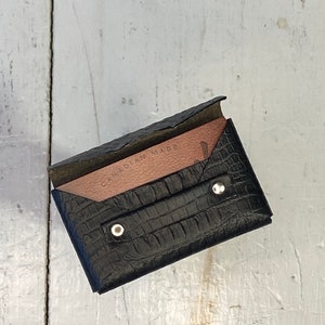 K&ouml;nnte beinhalten: Schwarzer Lederkartenhalter mit Krokodiltextur. Die Innenseite ist hellbraun und tr&auml;gt den Text "CANADIAN MADE". Der Kartenhalter wird mit zwei silbernen Druckkn&ouml;pfen verschlossen.
