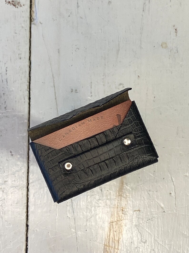 K&ouml;nnte beinhalten: Schwarzer Lederkartenhalter mit Krokodiltextur. Die Innenseite ist hellbraun und tr&auml;gt den Text "CANADIAN MADE". Der Kartenhalter wird mit zwei silbernen Druckkn&ouml;pfen verschlossen.