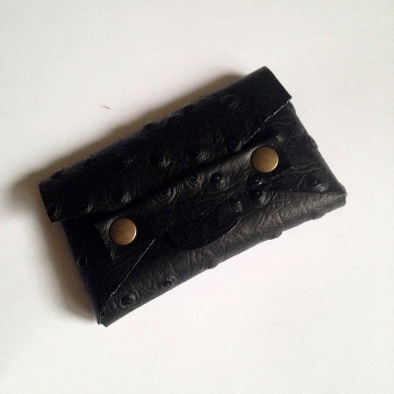 K&ouml;nnte beinhalten: Schwarzer Kartenhalter aus Leder mit strukturiertem Strau&szlig;enmuster. Die Geldb&ouml;rse hat einen Klappenverschluss, der mit zwei messingfarbenen Druckkn&ouml;pfen gesichert ist. Ein kleines, kompaktes Accessoire.