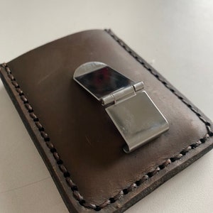 May include: A brown leather wallet with a metal money clip. The wallet has a stitched edge and is closed.