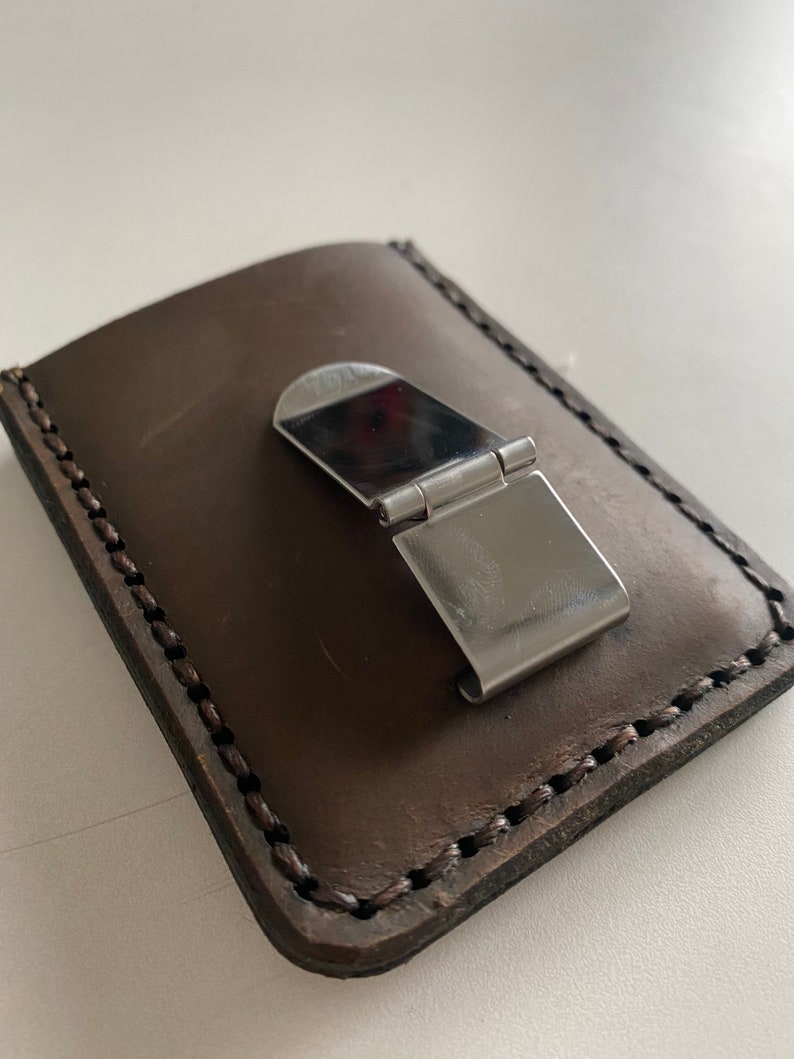 May include: A brown leather wallet with a metal money clip. The wallet has a stitched edge and is closed.