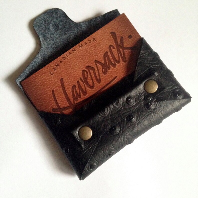 K&ouml;nnte beinhalten: Schwarzer Lederkartenhalter mit strukturierter Oberfl&auml;che und zwei messingfarbenen Druckkn&ouml;pfen. Eine braune Karte mit dem Wort "Haversack" und "Canadian Made" ist im Inneren sichtbar.