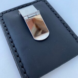 May include: A black leather money clip with a silver metal clip. The clip has a hinged design and a smooth, polished finish.