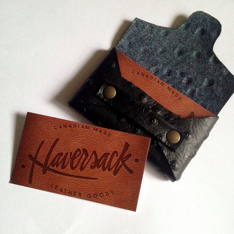 K&ouml;nnte beinhalten: Ein schwarzer Lederkartenhalter mit strukturierter Oberfl&auml;che und Druckknopfverschluss, neben einem braunen Lederetikett. Das Etikett tr&auml;gt die Aufschrift "CANADIAN MADE Haversack LEATHER GOODS". Die Innenseite des Kartenhalters ist aus braunem Leder.