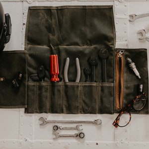 Peut inclure: Un sac à outils enroulable en toile verte avec une fermeture éclair et divers outils à l'intérieur, notamment des clés, des pinces, un tournevis et une bougie d'allumage. Le sac est posé sur une surface blanche avec un casque de moto, une tasse de café et un livre intitulé "Zen et l'art de l'entretien des motocyclettes" de Robert M. Pirsig.