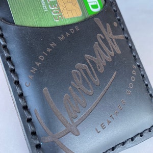 May include: Black leather wallet with a stitched edge and the text "Leather Goods" and "Canadian Made" printed on the front. The wallet has a slot for a credit card or SIM card.