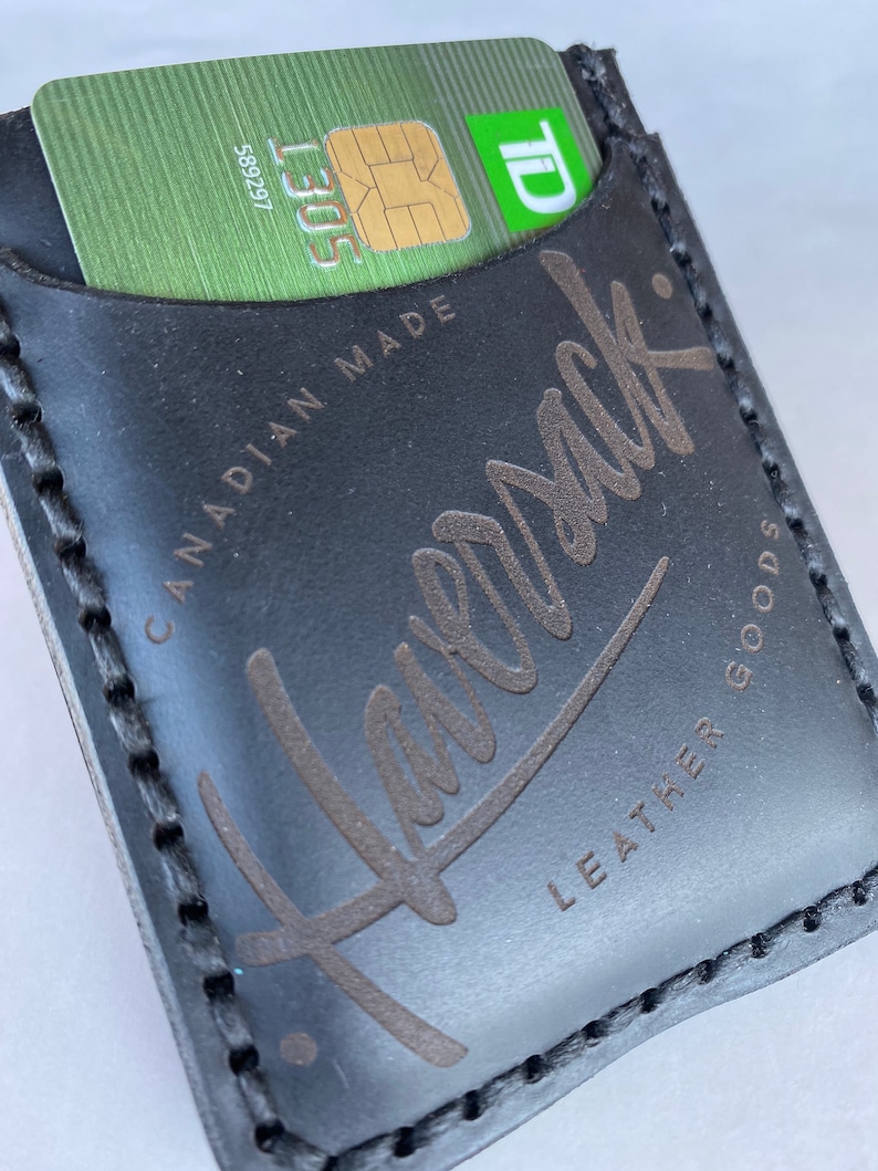May include: Black leather wallet with a stitched edge and the text "Leather Goods" and "Canadian Made" printed on the front. The wallet has a slot for a credit card or SIM card.