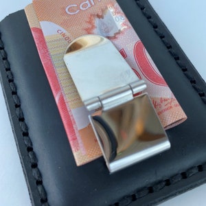 May include: A black leather wallet with a money clip. The money clip is made of metal and has a shiny finish. A Canadian five dollar bill is visible in the wallet.
