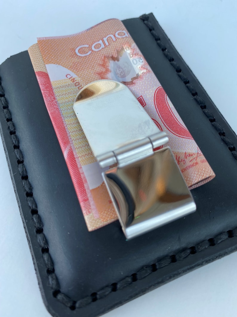 May include: A black leather wallet with a money clip. The money clip is made of metal and has a shiny finish. A Canadian five dollar bill is visible in the wallet.