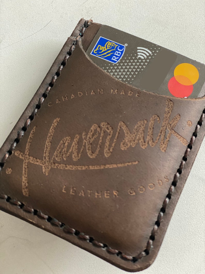 May include: A brown leather card holder with the text "Haversack Leather Goods" and "Canadian Made" embossed on the front. A credit card is visible in the pocket.