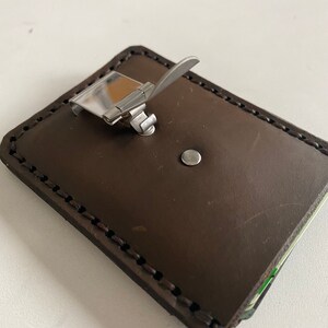 May include: A brown leather wallet with a metal clip closure. The wallet has a stitched edge and a small metal rivet.