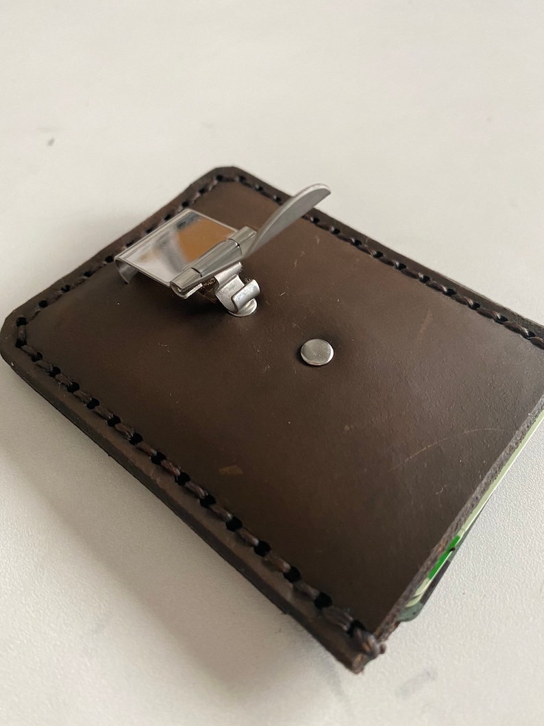 May include: A brown leather wallet with a metal clip closure. The wallet has a stitched edge and a small metal rivet.