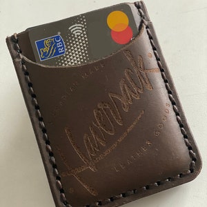 May include: A brown leather card holder with a stitched edge and the word "Remark" embossed on the front. The card holder is made in Canada and has the words "Leather Goods" printed on the bottom.