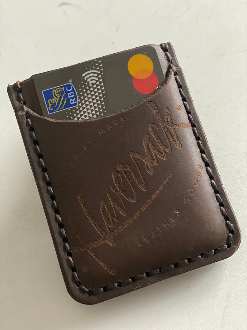 May include: A brown leather card holder with a stitched edge and the word "Remark" embossed on the front. The card holder is made in Canada and has the words "Leather Goods" printed on the bottom.