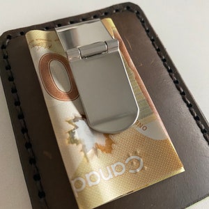 May include: A brown leather wallet with a silver money clip holding a Canadian dollar bill. The bill has the text "Canada" and "One" printed on it.