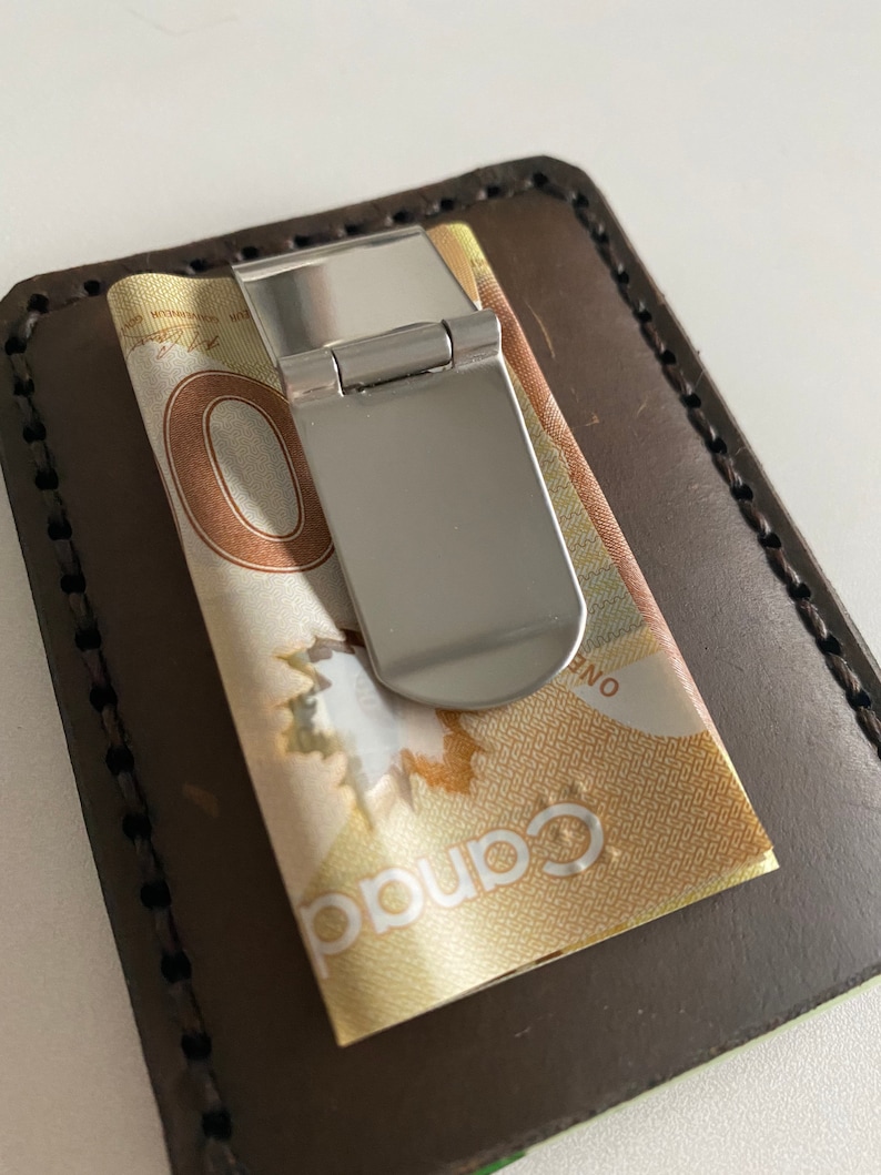 May include: A brown leather wallet with a silver money clip holding a Canadian dollar bill. The bill has the text "Canada" and "One" printed on it.