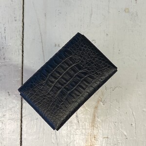 K&ouml;nnte beinhalten: Eine schwarze, rechteckige Geldb&ouml;rse mit einem strukturierten Muster in Krokodilleder-Optik. Die geschlossene Geldb&ouml;rse scheint aus Leder zu sein. Das Produkt wird auf einer wei&szlig;en, verwitterten Holzoberfl&auml;che pr&auml;sentiert.
