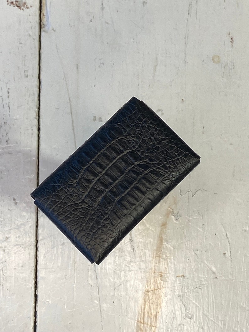 K&ouml;nnte beinhalten: Eine schwarze, rechteckige Geldb&ouml;rse mit einem strukturierten Muster in Krokodilleder-Optik. Die geschlossene Geldb&ouml;rse scheint aus Leder zu sein. Das Produkt wird auf einer wei&szlig;en, verwitterten Holzoberfl&auml;che pr&auml;sentiert.
