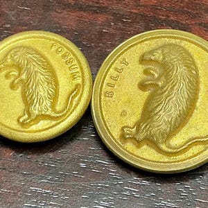 May include: Two gold-colored, round metal buttons. Each button features a detailed relief of a possum and the text "BILLY POSSUM" around the edge. The buttons are displayed on a dark brown surface.