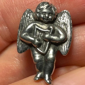 Könnte beinhalten: Kleine, silberfarbene Engelsfigur mit detaillierten Flügeln und einer Harfe. Der Engel ist in stehender Position mit einem lächelnden Gesichtsausdruck dargestellt. Die Figur ist wahrscheinlich aus Metall und hat ein Vintage- oder antikes Aussehen.