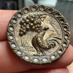 May include: Antique brass button with a detailed relief of a rooster, grapes, and foliage. The button has a decorative border with a series of small, raised dots. The rooster is facing left, with its tail feathers fanned out. A vintage collectible.