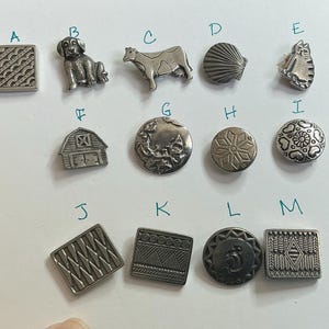 Könnte beinhalten: Eine Sammlung von silberfarbenen Metallknöpfen mit verschiedenen Designs. Die Knöpfe zeigen Bilder von einem Hund, einer Kuh, einer Muschel, einer Katze, einer Scheune und anderen dekorativen Mustern. Die Knöpfe sind auf einer weißen Oberfläche angeordnet.
