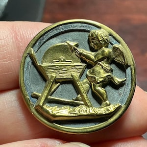 May include: A round, antique button with a gold-colored cherub figure sharpening a blade on a grindstone. The cherub has wings and is wearing a short tunic. The button has a dark gray background with a gold border.