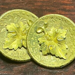 Puede incluir: Dos botones antiguos de latón. Cada botón es redondo con un diseño de hoja en relieve en el centro. Los botones tienen un fondo texturizado con un patrón floral. Los botones son de color dorado.