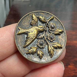 May include: A round, antique brass button with a detailed design. A gold-colored bird is depicted in flight, holding a sprig of leaves and flowers. The button has a textured, aged appearance, with a decorative rope-like edge.