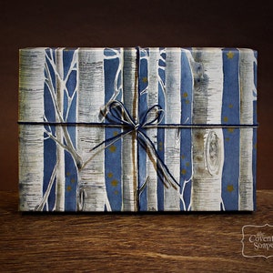 May include: A rectangular gift box wrapped in paper with a birch tree design in white, blue, and silver. The box is tied with navy blue twine and a bow. Gold stars are scattered across the blue stripes.