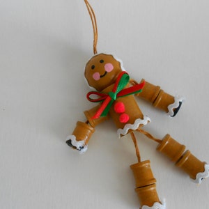 Puede incluir: Un adorno navideño de hombre de jengibre hecho de madera pintada de marrón con detalles de glaseado blanco. El adorno tiene un lazo rojo y verde y puntos rojos pintados en el cuerpo. El adorno está encordado con un cordel.