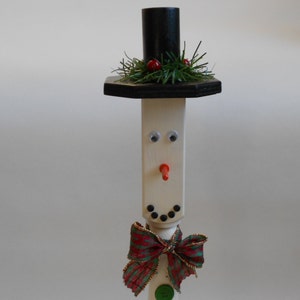 May include: A white wooden snowman figurine with a black top hat, red berries, and a green and red bow. The snowman has black eyes, a red nose, and a black mouth.
