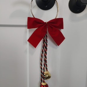 May include: A door hanger featuring a large red velvet bow, gold ring, and decorative braided cords in black, red, and gold. The cords are adorned with three bells in gold, red, and green. The door hanger is designed for holiday or seasonal decor.
