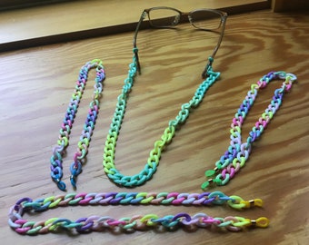 May include: Four colorful plastic chains for glasses. The chains are in shades of pink, purple, blue, green, and yellow. The chains are laid out on a wooden surface.