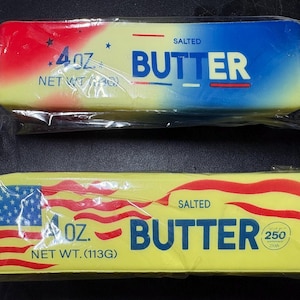 Puede incluir: Dos barras rectangulares de mantequilla salada en envoltorios de plástico transparente. La barra superior presenta un degradado rojo, blanco y azul con estrellas y el texto 4 OZ. NET WT. 113G. La barra inferior tiene un fondo amarillo con un diseño de bandera roja y blanca, el texto 4 OZ. NET WT. 113G y un emblema del 250 aniversario.