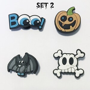 Op de afbeelding: Set van vier Halloween-schoenbedels. De bedels zijn een pompoen met een glimlachend gezicht, een vleermuis met blauwe voeten, een schedel met kruisbeenderen en een blauwe en witte "Boo!" bedel met oogjes.